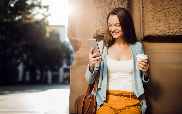 Frau hält ein Smartphone in der einen und einen Kaffeebecher in der anderen Hand