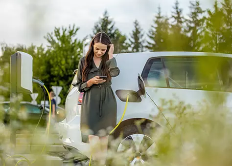 Frau schaut auf ein Smartphone, im Hintergrund lädt ein Elektroauto