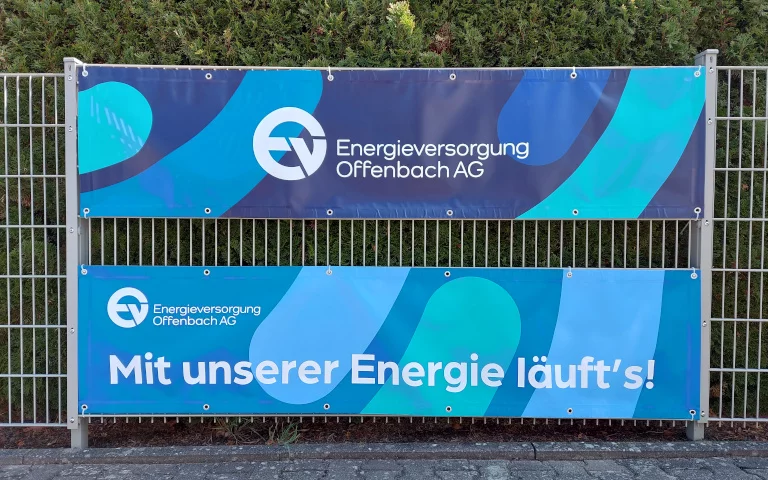 Zwei EVO-Banner an einem Metallzaun mit Logo der Energieversorgung Offenbach AG und dem Slogan ‚Mit unserer Energie läuft’s!‘ auf blau-grünem Hintergrund.